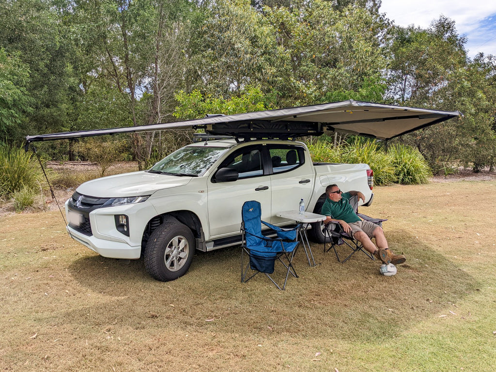 OUTBOUND Shield 3 Freestanding Awning open mounted on white dual cab Ute. There is a table and two chairs next to the Ute with a man sitting in one of the chairs. Set on grass with trees in the background.